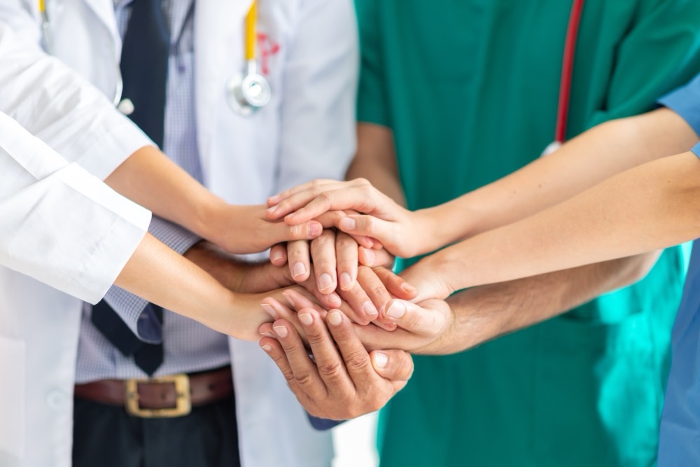 A group of healthcare professionals stack their hands together in a show of teamwork, wearing white coats, scrubs, and stethoscopes.
