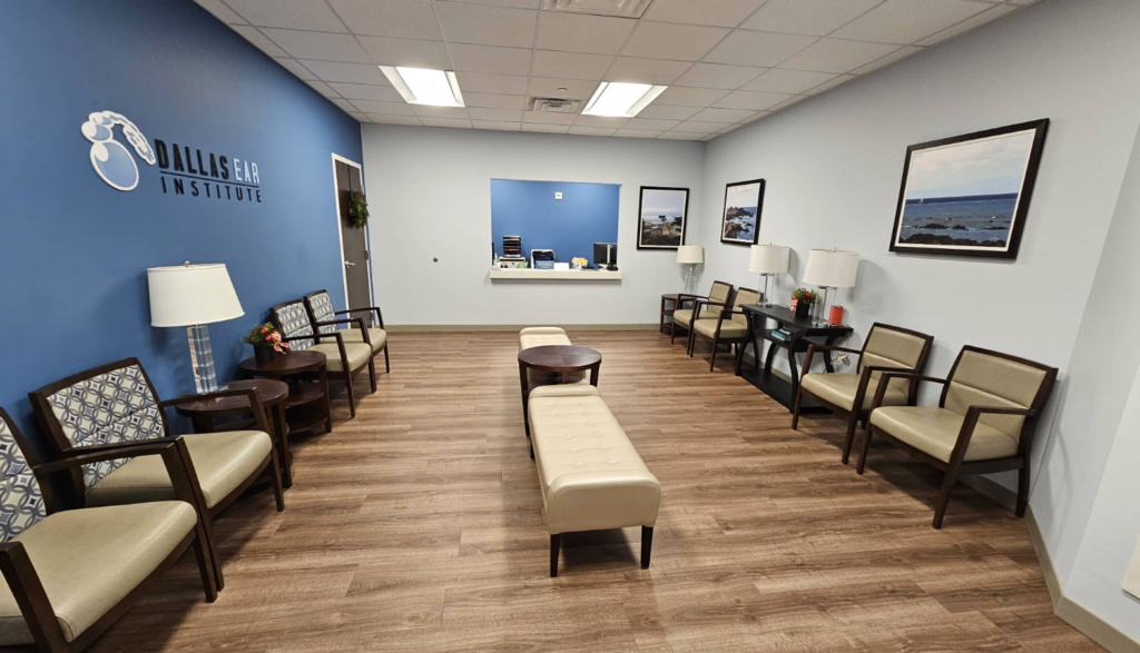 A medical office waiting room with tan chairs, two benches, side tables with lamps, and wall art. The Dallas Ear Institute logo is on the blue wall near the reception window. The floor is wood-patterned.